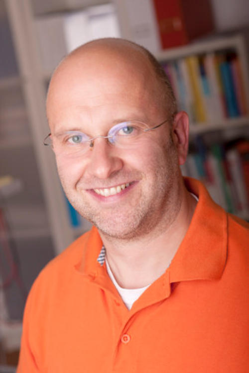 Porträt von Heilpraktiker Olaf Preuss mit Brille und orangefarbenem Poloshirt vor einem Bücherregal